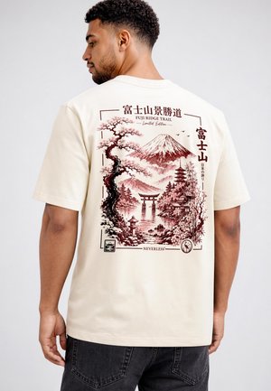 Man wearing off-white t-shirt with maroon Japanese landscape design showing Mount Fuji, cherry blossoms, pagoda, and torii gate on back.