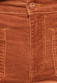 Pantalones de pana en color óxido con estrías verticales, que cuentan con dos bolsillos frontales y un botón marrón de marca con costura amarilla.