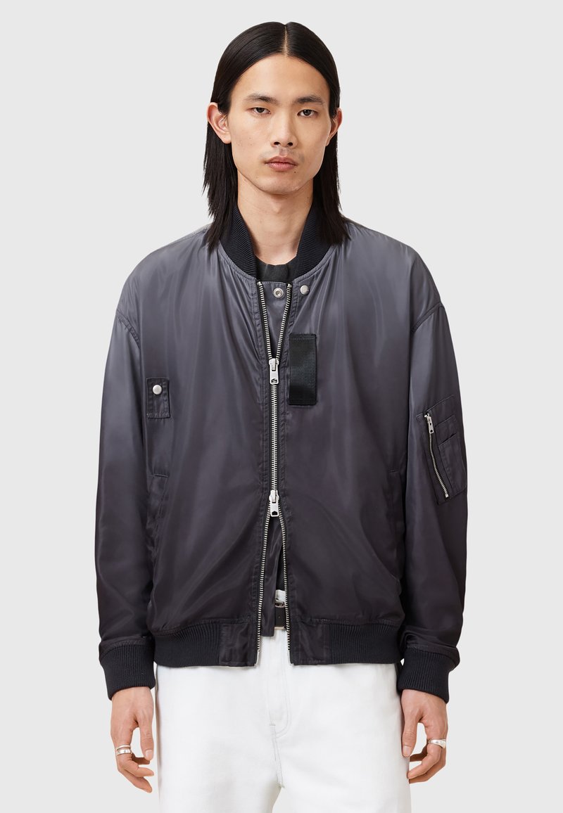 Veste bomber noire légère avec une finition brillante, des poignets côtelés, une fermeture éclair et plusieurs poches. Texture lisse avec un minimalisme dans les détails.