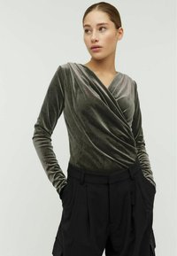 Fluwelen top in een wikkelstijl in donkergroen met lange mouwen, met een gedrapeerde halslijn. Gecombineerd met hooggeplaatste zwarte broek.