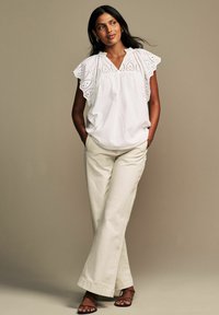 Blusa bianca con dettagli in pizzo e maniche a volant, abbinata a pantaloni beige a gamba larga e sandali marroni con lacci. Tessuto morbido e leggero.