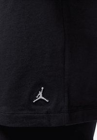 Black cotton T-shirt with a rounded hem. Features a white embroidered Jumpman logo on the lower left side. Smooth texture, short sleeve design.
