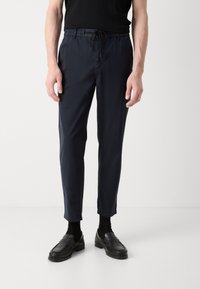 Mörkblå tapered byxor med elastiskt midjeband och dragsko, tillverkade av ett slätt tyg. Kombinerat med svarta loafers och strumpor.