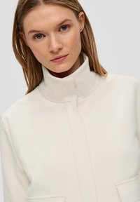 Crèmegekleurde jas met hoge kraag, gemaakt van zachte stof, met een sluiting aan de voorkant en twee zijzakken. Glad oppervlak, minimalistisch ontwerp.