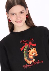 Sudadera negra con un gráfico colorido de un oso de peluche dentro de una corona, con texto rojo que dice “Feliz Navidad”. Pendientes de cinta roja.