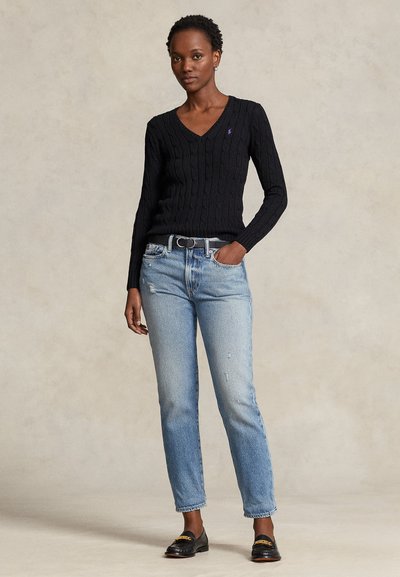 Ženska stoji z roko v žepu, oblečena v črni pulover z V-izrezom in kabelskim vzorcem, svetlo modre kavbojke, črni pas in črne mokasine.