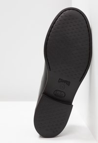 Semelle de chaussure noire avec grip texturé, marquée "Camper" et talon en caoutchouc, présentée sur fond blanc et surface d'angle verticale.