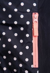 Tessuto blu marina con pois bianchi e zip rosa. Texture liscia con una finitura lucida, che mette in evidenza un dettaglio della zip a contrasto.