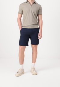 Polo à manches courtes zippé pour homme en gris clair, associé à un short marine foncé et à des baskets couleur crème avec des lacets.