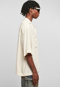 Chemise à manches courtes couleur beige clair avec une poche, coupe ample et texture lisse, associée à un jean bleu déchiré et des lunettes de soleil.