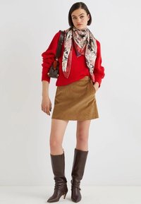 Maglione rosso a maglia, gonna in velluto a coste marrone, sciarpa di seta a fantasia, borsa a tracolla in pelle marrone, stivali marroni alti fino al ginocchio con punta affilata.