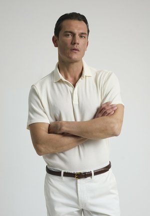 Camisa polo de manga corta color crema con tejido texturizado, combinada con pantalones blancos y un cinturón de cuero marrón. El modelo está de pie con los brazos cruzados.