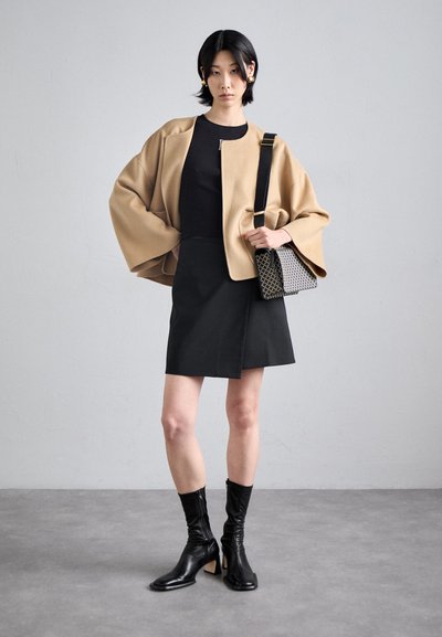 Giacca oversize in misto lana beige, top aderente nero, gonna avvolgente nera, stivali in pelle neri alla caviglia e una piccola borsa a tracolla con motivo.