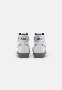 Bílé vysoké tenisky s texturovaným koženým svrškem, šedou podešví a výrazným černým nápisem "NIKE" na zadní straně.