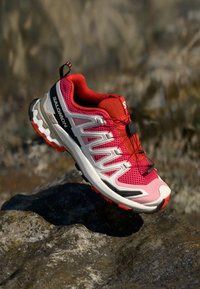 Scarpa sportiva rosa e grigia con tomaia in rete testurizzata, suola in gomma e accenti neri. Presenta un design con lacci e un tab più lungo sul retro.