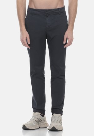 Uomo con pantaloni aderenti grigio scuro e sneakers New Balance di colore chiaro, in piedi contro uno sfondo bianco uniforme.