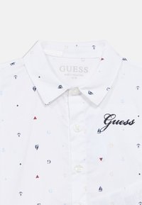 Valkoinen nappipaita, jossa on merihenkinen kuvio, jossa pieniä veneitä, ankkureita ja geometrisia muotoja sinisen, punaisen ja beigen sävyissä. "Guess"-logo brodeerattu.