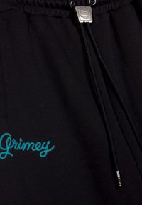 Los pantalones de chándal negros presentan un cordón con un extremo de metal plateado y bordado en turquesa que dice "Grimey" en la pierna. Textura de suave tela de algodón.