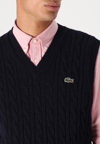 Ναυτικό πλεχτό γιλέκο με V-λαιμόκοψη; υφή με σχέδια; διαθέτει κεντητό λογότυπο κροκόδειλου σε πράσινο χρώμα. Κάτω από αυτό υπάρχει ένα ανοιχτό ροζ πουκάμισο με ρίγες.