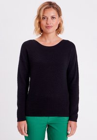 Pull noir texturé avec un col rond et des manches longues, associé à un pantalon vert. Le tricot présente un motif subtil et une coupe décontractée.