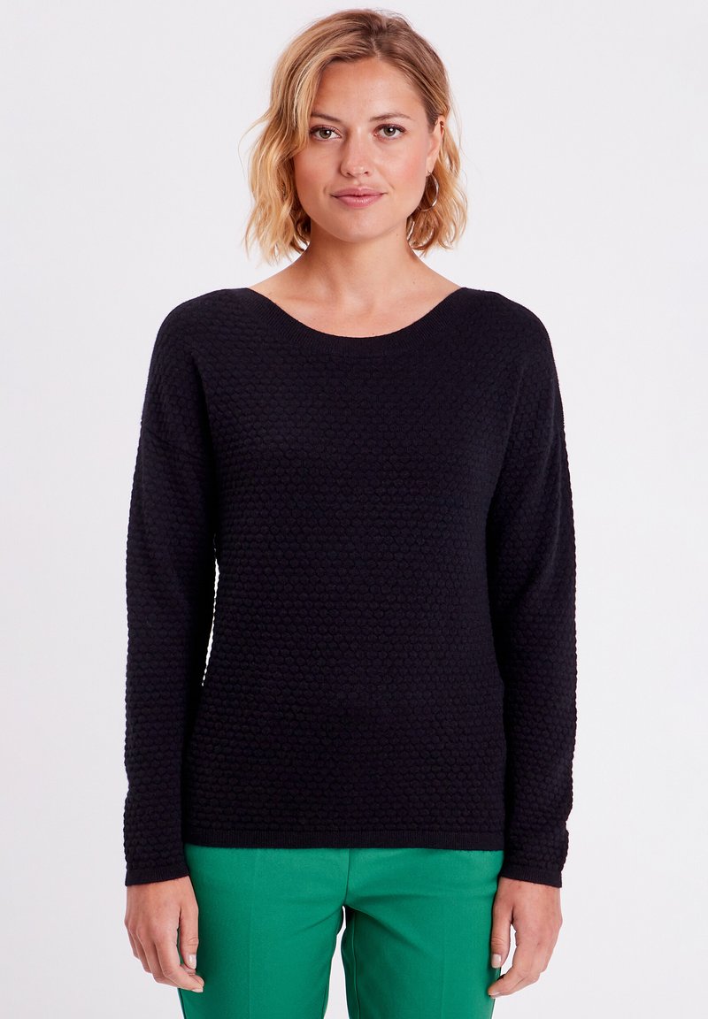 Pull noir texturé avec un col rond et des manches longues, associé à un pantalon vert. Le tricot présente un motif subtil et une coupe décontractée.