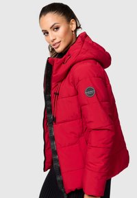 Red padded jacket with a hood, zip closure, textured fabric, black accents, and a logo patch on the sleeve. Smooth, lightweight design.