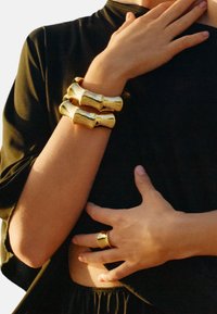 Guldtonat smycke som innehåller två breda, texturerade armband och en geometrisk ring, med en glänsande yta och släta konturer.