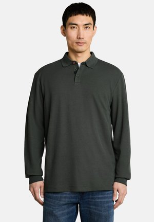 Homme aux cheveux courts et noirs portant un polo vert foncé à manches longues et un jean bleu sur un fond clair uni.