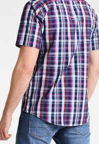 Man med kortärmad rutiga skjorta i röd, vit och blå, tillsammans med blå jeans, sedd bakifrån mot en vit bakgrund.