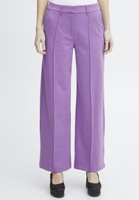 Pantalons larges violets avec une texture lisse, dotés de plis à l'avant et de poches latérales, assortis à des chaussures à talons hauts noires.