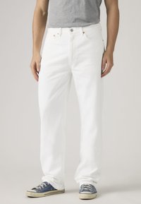 Άτομο που φοράει χαλαρά λευκά τζιν με μπροστινές τσέπες και κουμπωτό κλείσιμο, συνδυασμένα με μπλε πάνινα αθλητικά παπούτσια και γκρι μπλουζάκι που έχει μπει μέσα στο παντελόνι.