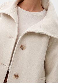 Beige kabát z vlněné směsi s širokým límcem, dvěma knoflíky a texturovaným povrchem. Pod ním je světle béžová košile.