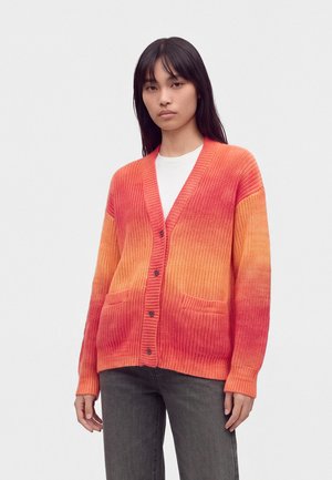 Mujer que lleva un cárdigan degradado naranja y rojo, camiseta blanca, vaqueros anchos de pierna oscura y zapatillas rojas y beige, de pie sobre un fondo blanco.