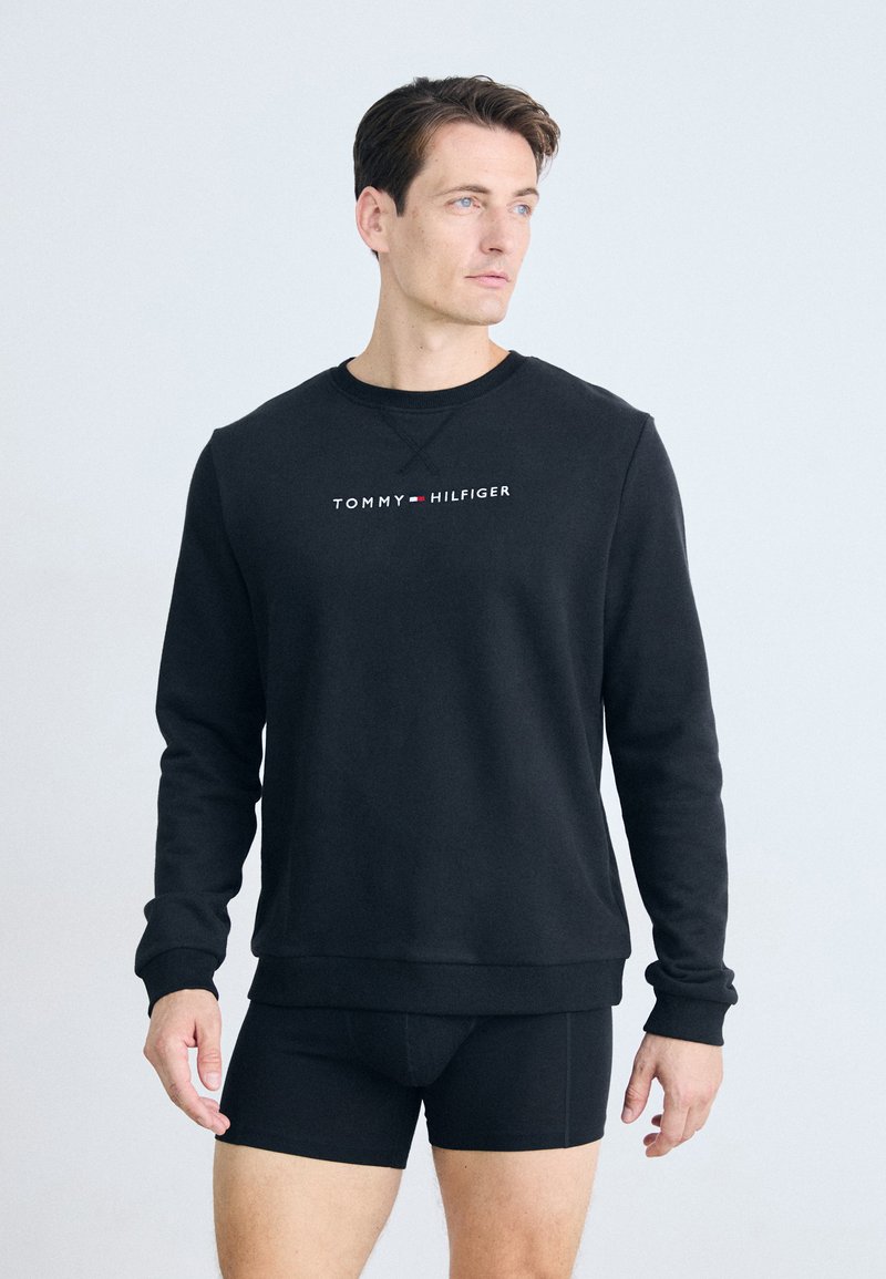 Sudadera negra con cuello redondo y mangas largas, presenta un logo en rojo y blanco de "TOMMY HILFIGER" en el pecho. Textura de tela suave.