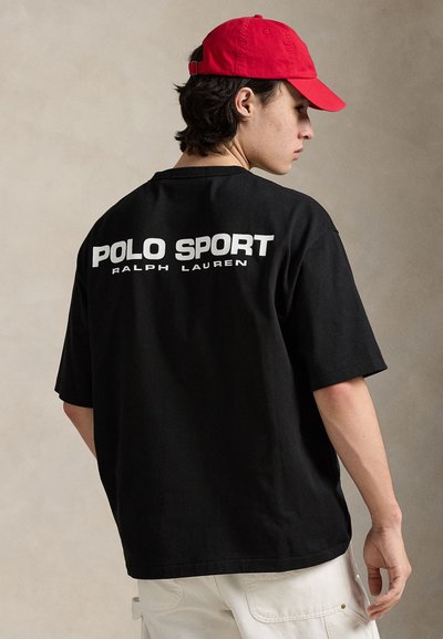 Μαύρο κοντομάνικο t-shirt με μεγάλο λευκό λογότυπο "POLO SPORT RALPH LAUREN" στην πλάτη. Φορεμένο με κόκκινο καπέλο.
