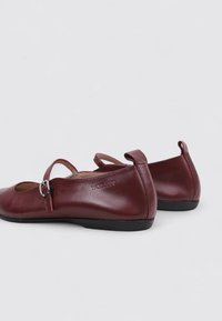 Zapatos Mary Jane de cuero rojo oscuro con una pequeña correa de hebilla, puntera redondeada y suela plana negra sobre un fondo claro y liso.