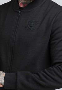 Giacca bomber nera con zip in misto cotone, caratterizzata da colletto e polsini a coste, e un logo ricamato discreto sul petto.
