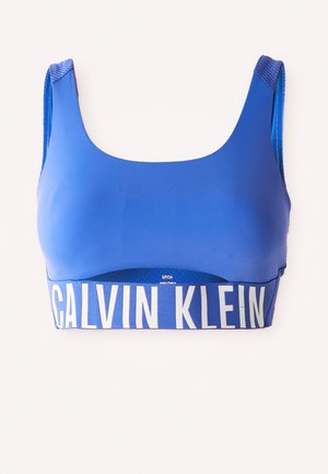 Blauwe sportbeha met brede banden en een grote witte 'Calvin Klein' logo band aan de onderkant, weergegeven tegen een witte achtergrond.