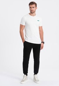 Bílý tričko s crew neck výstřihem a malým tyrkysovým akcentem, v kombinaci s černými tepláky. Model má na sobě světle šedé tenisky a stojí na jednobarevném pozadí.