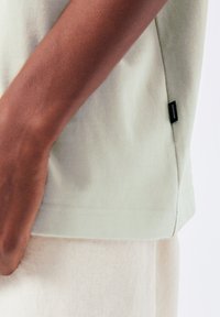Light green cotton T-shirt with short sleeves, side slit, and a small black brand label near the hem. Background is plain white.