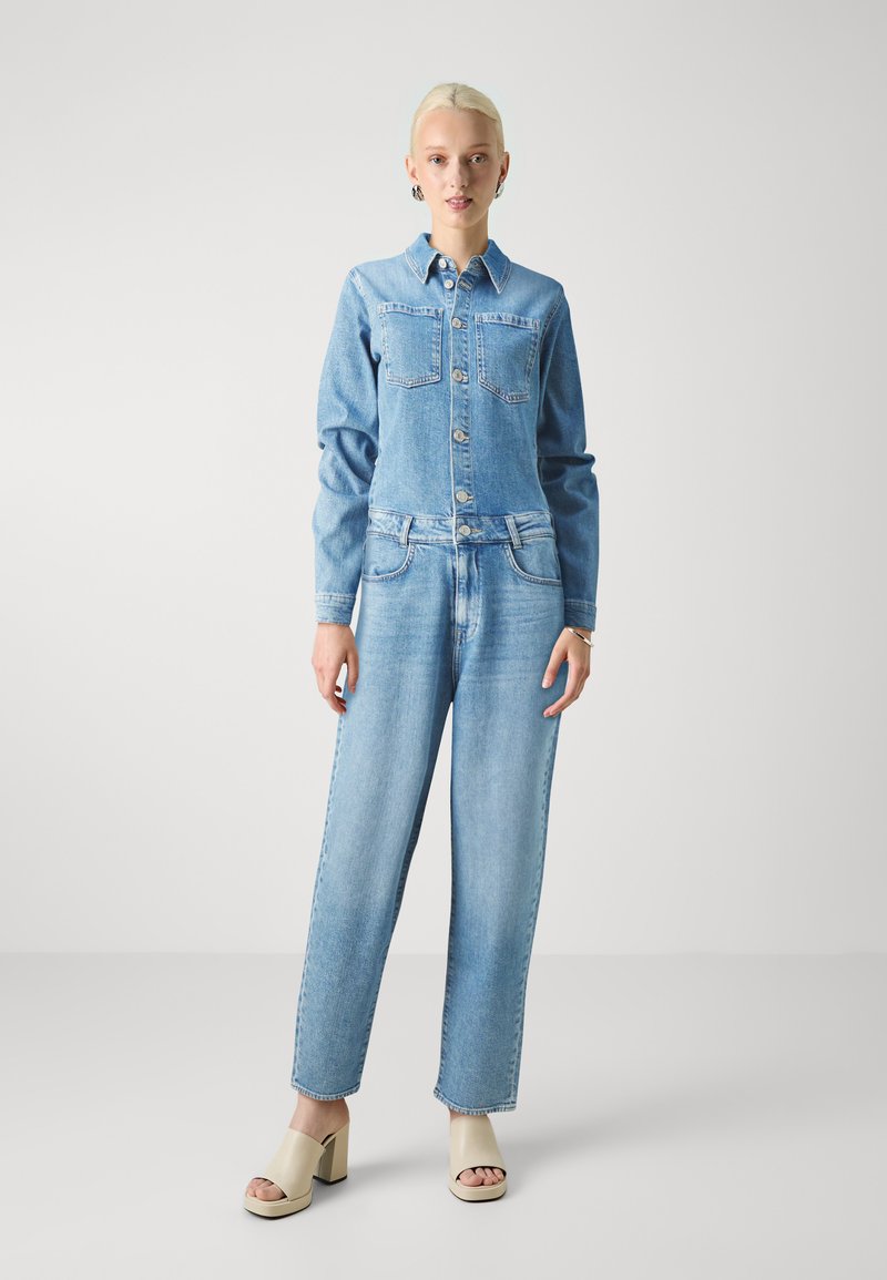 Light blue denim jumpsuit with long sleeves, front button closure, two chest pockets, and straight-leg fit, paired with beige platform sandals.