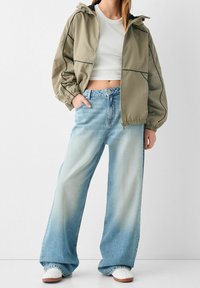Osoba ubrana w jasnozieloną kurtkę z kapturem, białą krótką bluzkę, szerokie wyblakłe niebieskie dżinsy i białe trampki na jednolitym tle.