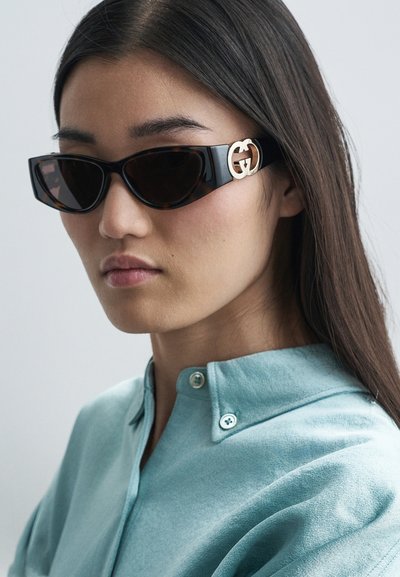 Juodos katės akių saulės akiniai su auksiniu GG logotipu ant šventyklų. Lygi, blizgi apdaila; tamsiai tonuoti objektyvai; modelis vilki šviesiai mėlyną apykaklę.