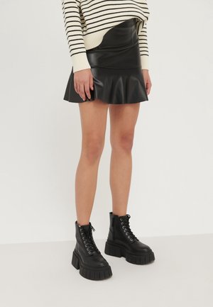 Donna che indossa una minigonna in pelle nera con bordo arricciato, stivali platform neri con lacci, e un maglione a righe crema e nere.
