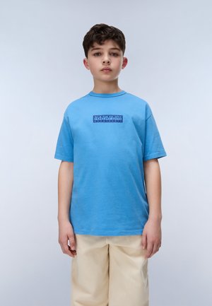 Jongen met kort donker haar, een blauwe Napapijri T-shirt en beige broek dragend, staande tegen een eenvoudige lichte achtergrond.