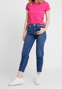 Rosa Polohemd mit Kragen und Logo, kombiniert mit blauen hochgeschnittenen Jeans. Weiße Sneakers mit strukturiertem Sohlen. Einfaches, lässiges Outfit.
