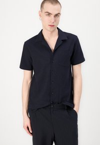 Camicia navy a maniche corte con bottoni in tessuto testurizzato, dotata di taschino e colletto rilassato, abbinata a pantaloni scuri a righe.