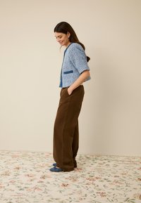 Hellblaue Jacke mit Struktur und kurzen Ärmeln, dunkelblaue Taschenapplikationen, kombiniert mit braunen, weit geschnittenen Hosen und blauen Schuhen.