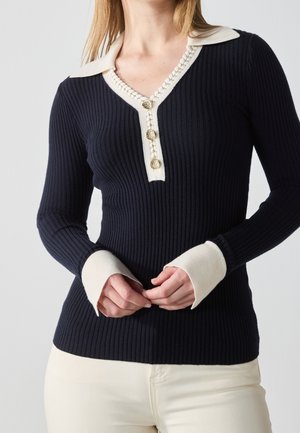 Kobieta w dopasowanym granatowym swetrze w prążki z kremowym kołnierzem, mankietami i trzema złotymi dekoracyjnymi guzikami przy dekolcie, w połączeniu z jasnymi spodniami.