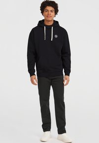 Sweat à capuche noir avec cordons blancs, poche avant et logo circulaire. Porté avec un pantalon sombre et des baskets blanches sur un fond neutre.